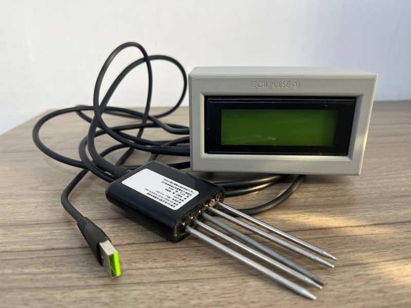 The SoilPulse Probe: a grey reader with a green screen, connected by cable to a black soil probe with five stainless steel rods
